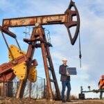 Oil worker in formal attire inspecting an oil pumpjack with a tablet.