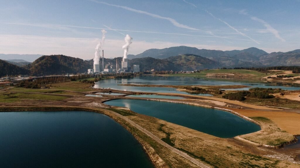 "Aerial view of an industrial power plant emitting smoke, surrounded by lakes and mountains.