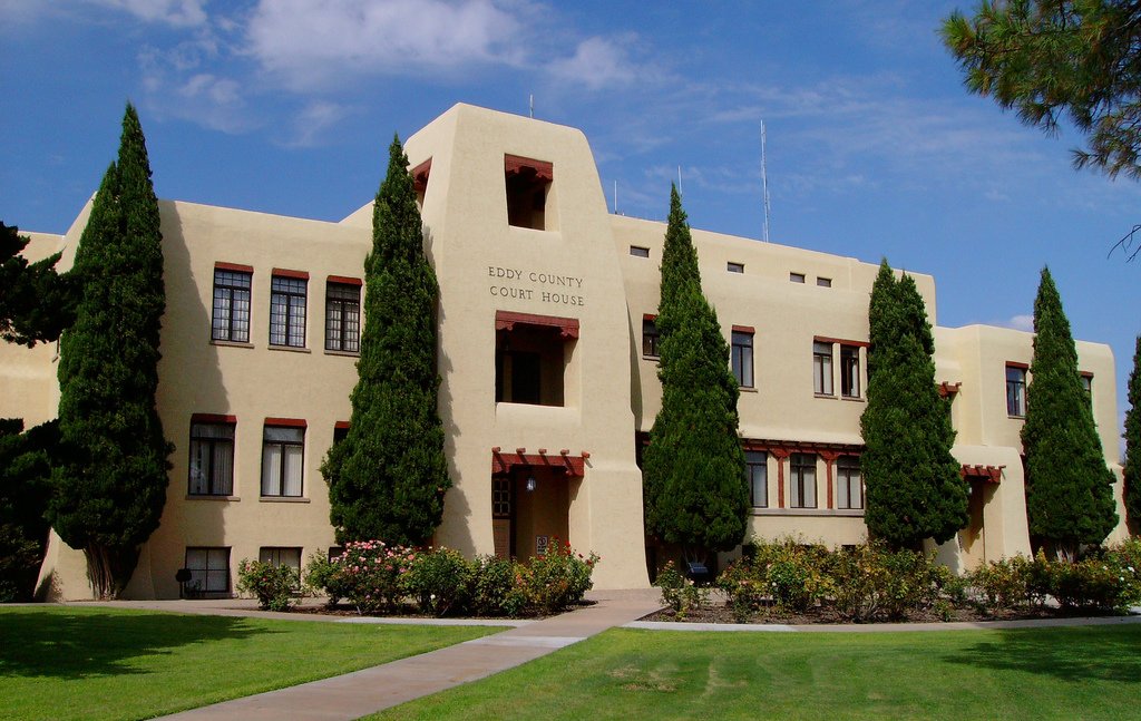 Eddy County Courthouse with classic Pueblo Revival architecture, stucco walls, and tall cypress trees.