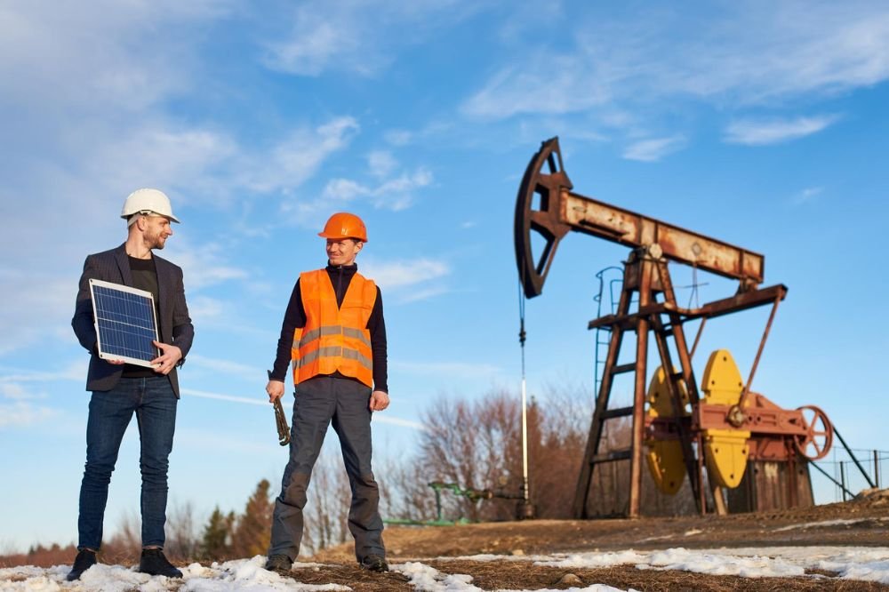 Two workers discussing renewable energy and oil extraction, with one holding a solar panel and the other wearing an orange safety vest near an oil pump jack.