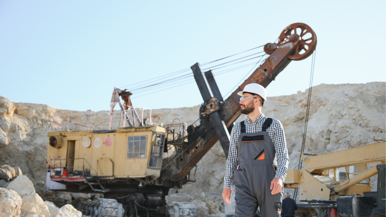 Worker in safety gear walking through an open-pit mining site with a large excavation machine in the background.