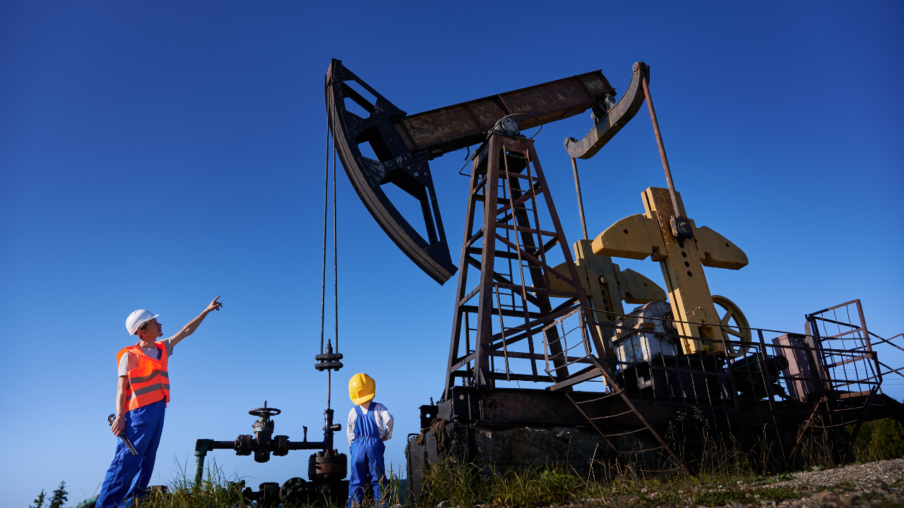 Oil field workers, including a child in safety gear, examining a pumpjack under a bright blue sky.